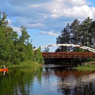 This image shows a bridge over a lake narrows in northern Minnesota. The Mesabi Trail paved cycling trail crosses the bridge and a canoe is in the lake.