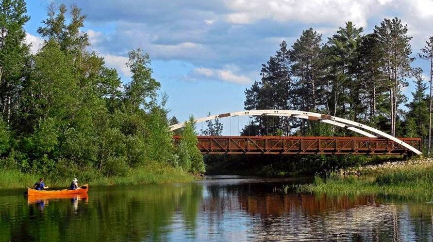 This image shows a bridge over a lake narrows in northern Minnesota. The Mesabi Trail paved cycling trail crosses the bridge and a canoe is in the lake.