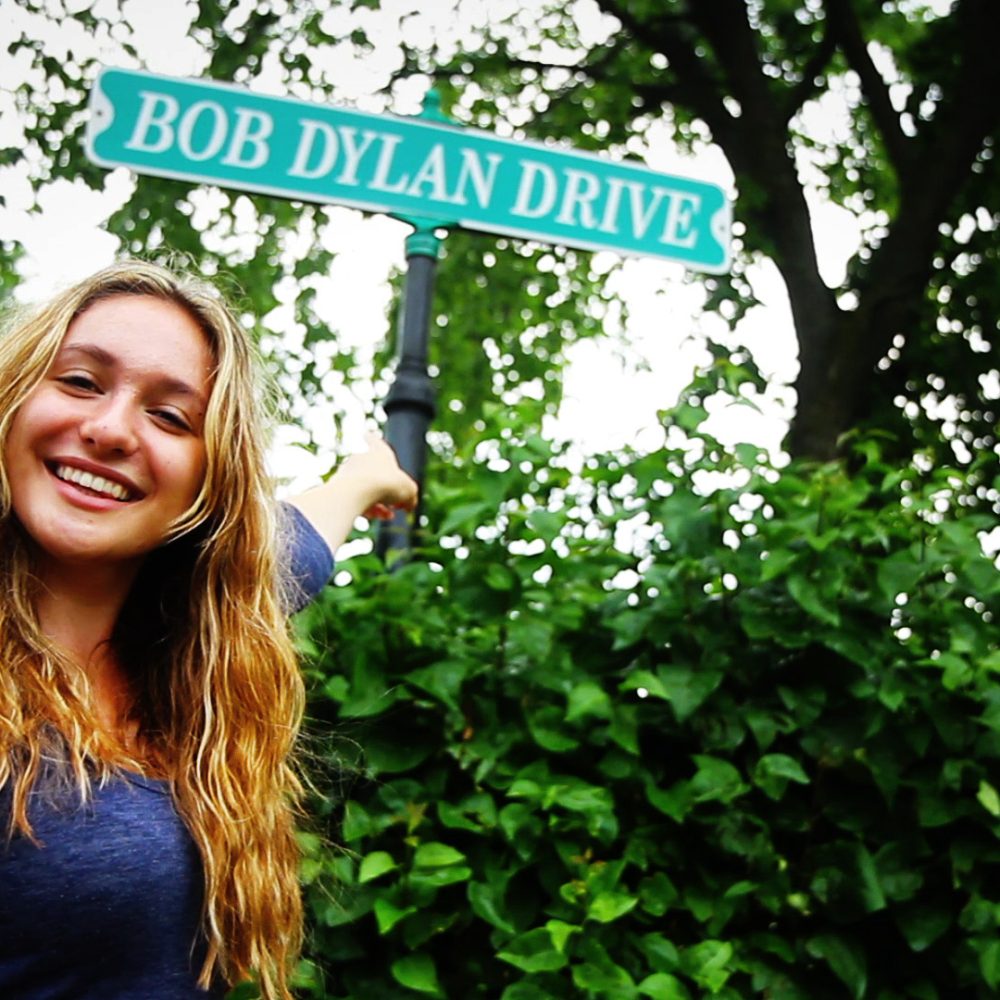 A young woman smiles as she points to the Bob Dylan Drive street sign in Hibbing, MN