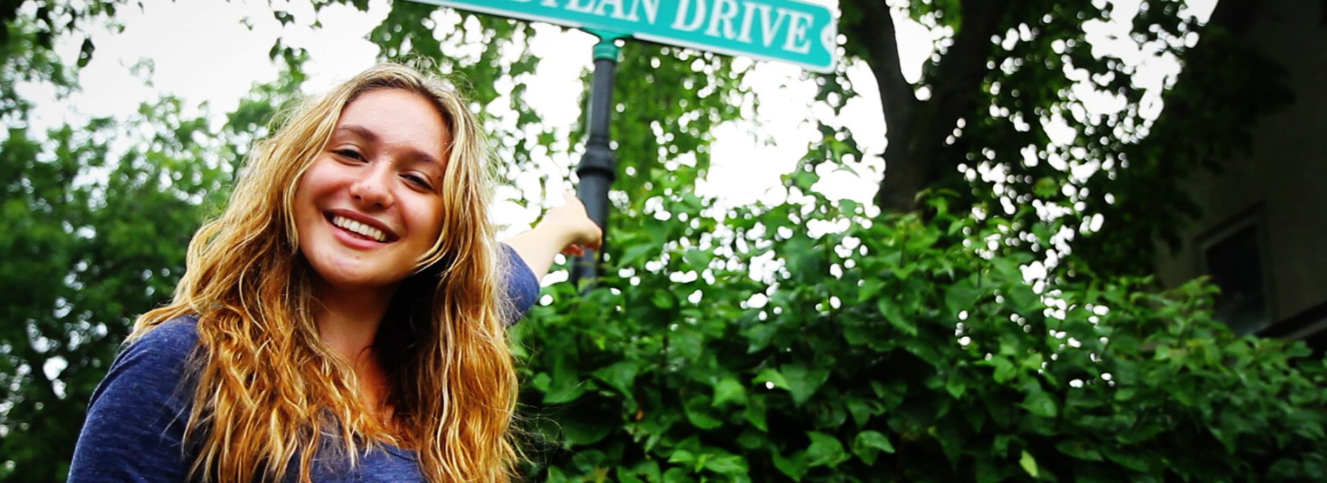 A young woman smiles as she points to the Bob Dylan Drive street sign in Hibbing, MN