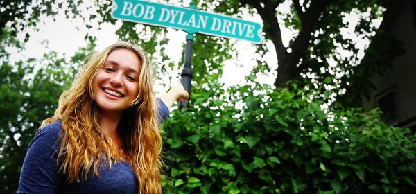 A young woman smiles as she points to the Bob Dylan Drive street sign in Hibbing, MN