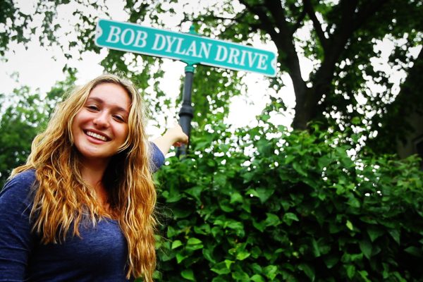 A young woman smiles as she points to the Bob Dylan Drive street sign in Hibbing, MN