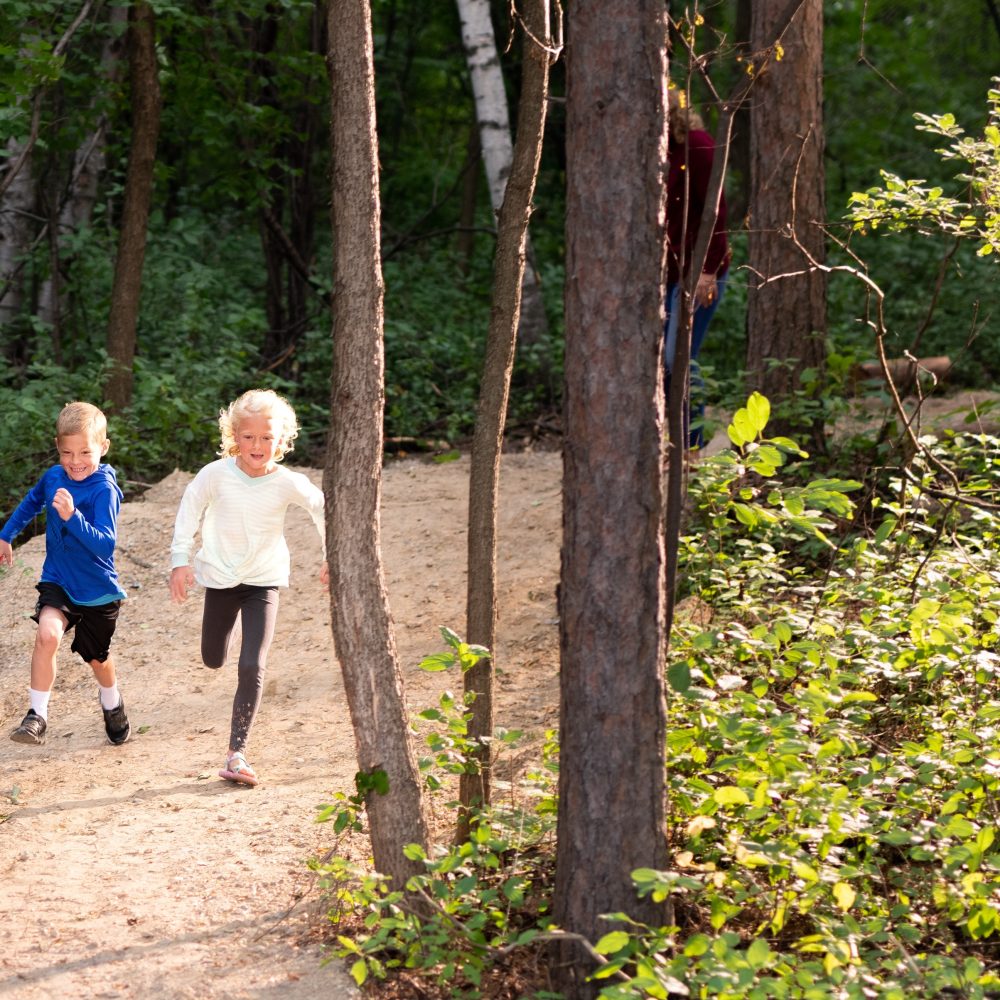 Two young children run joyfully among tall trees on a trail in northern Minnesota.