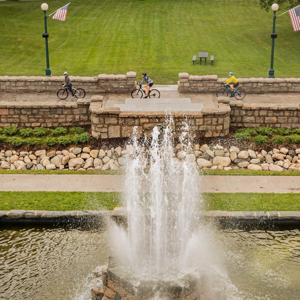 Three cyclists ride next to a fountain in Olcott Park, Virginia, MN