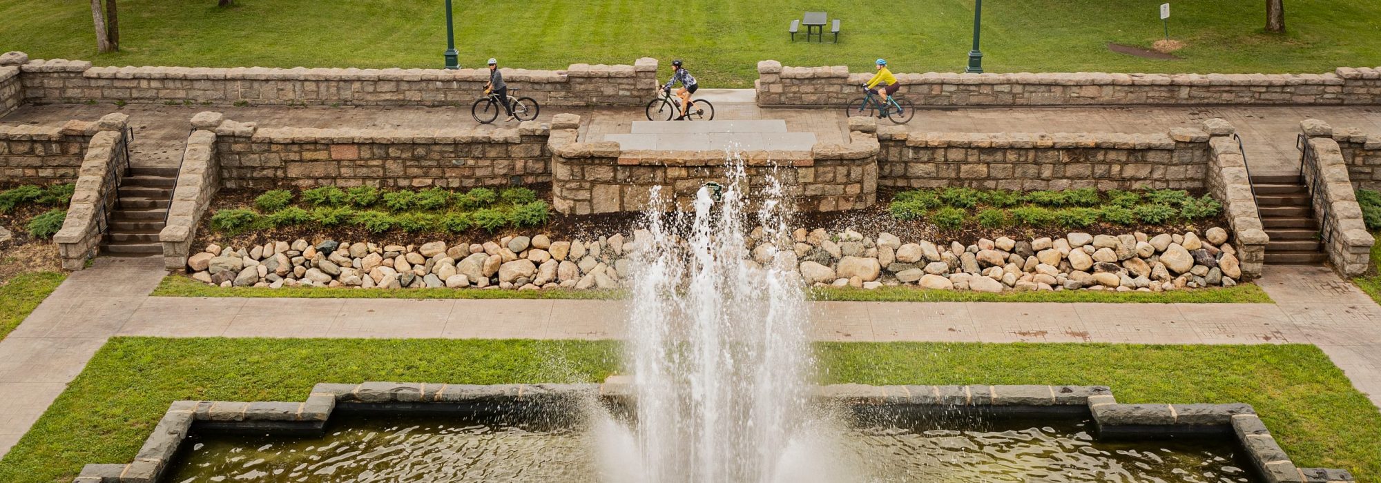 Three cyclists ride next to a fountain in Olcott Park, Virginia, MN