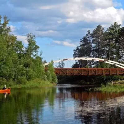 This image shows a bridge over a lake narrows in northern Minnesota. The Mesabi Trail paved cycling trail crosses the bridge and a canoe is in the lake.