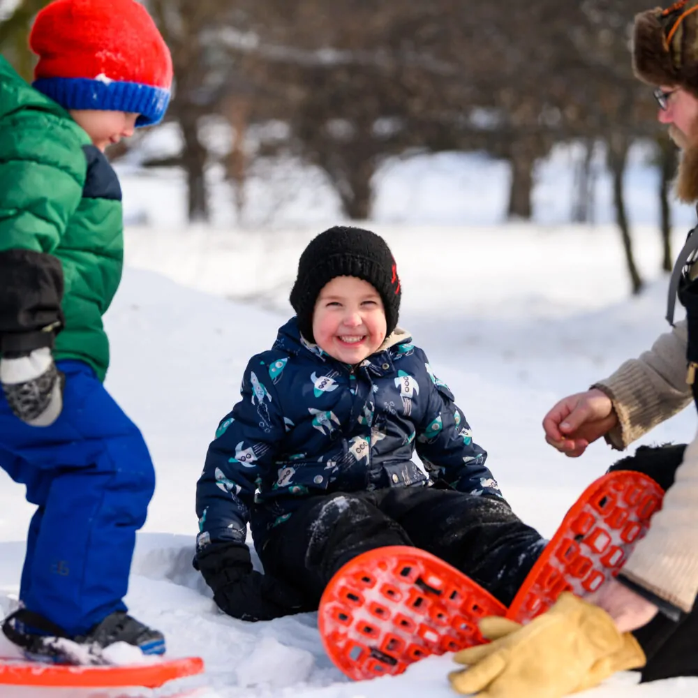 A little boy smiles as his dad puts snowshoes on him in northern Minnesota at Minnesota Discovery Center in Chisholm